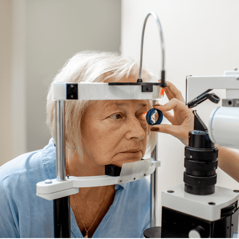 Photograph of elderly woman during an optometric exam, with her chin in a chinrest. There is a light shining in her eye as she is being examined by the doctor.